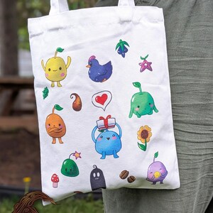 May include: White tote bag with colourful cartoon illustrations of various creatures and objects, including a yellow fruit-like character, a blue bird, a heart, and a gift. The bag is held by a person wearing green trousers.