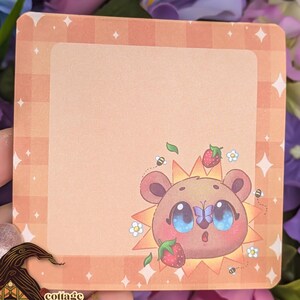 May include: Square-shaped sticker with a cartoon bear illustration. The bear has large blue eyes, a surprised expression, and is surrounded by a sunburst design. Strawberries and bees are also featured. The sticker has a peach-colored background with a border.