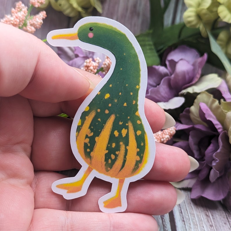 May include: A green and yellow gourd-shaped duck sticker with a white outline. The duck has a yellow beak and feet and a small pink blush on its cheek.
