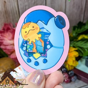 May include: A sticker featuring a cartoon-style illustration of a blue and pink virtual pet device with a yellow creature inside. The sticker has a pink border and is set against a blurred background with flowers.