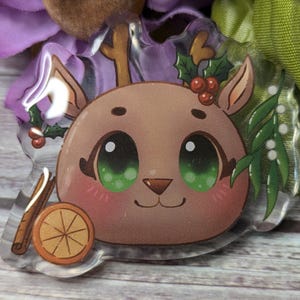 May include: A clear acrylic charm featuring a cartoon reindeer with large green eyes, a brown nose, and a red holly sprig with berries on its head. The reindeer is surrounded by green leaves and a cinnamon stick.