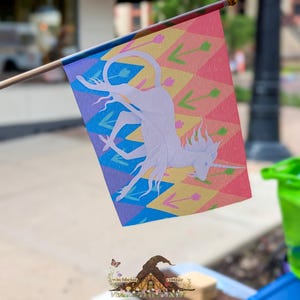 May include: A rectangular flag attached to a wooden pole featuring a light purple unicorn silhouette centered over a background of colorful diamond shapes in shades of blue, yellow, and pink, accented with small green arrow and pink flower icons.