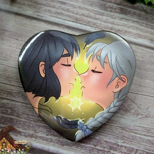 May include: Heart-shaped button featuring an illustration of two anime-style characters kissing, with closed eyes and rosy cheeks. The background has a yellow glow. The button has a glossy finish and is set against a wooden surface.