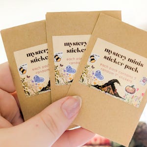 May include: Three small, brown paper packs labeled "mystery sticker" and "mystery minis sticker pack" are held in a hand. Each pack contains random 1" stickers. The stickers feature illustrations of a bee, a ghost, a witch's hat, and a pumpkin.