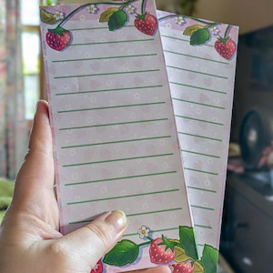 May include: Two pink notepads with a strawberry design. Each notepad has a border of strawberries, green leaves, and white flowers. The notepads have horizontal lines for writing. The background is a light pink color with small strawberry illustrations.