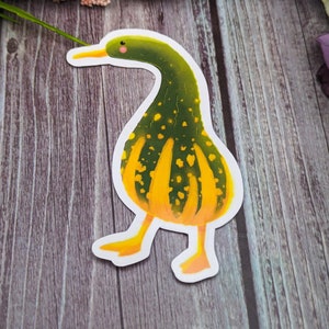 May include: A green and yellow gourd shaped like a duck with a white outline. The gourd has a yellow beak and a small pink blush on its cheek.