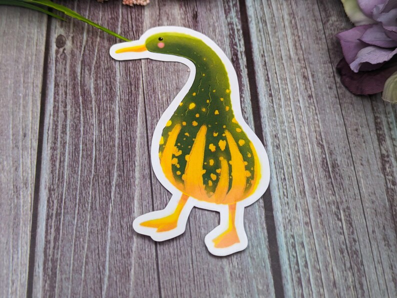 May include: A green and yellow gourd shaped like a duck with a white outline. The gourd has a yellow beak and a small pink blush on its cheek.