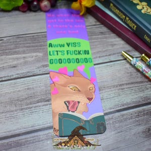May include: A rectangular paper bookmark featuring a purple and green background with the text Me when I get to the top & there's only one bed and Aww YISS LET'S FUCKIN GOOOOOOOOO. The design includes a tan cat with pink hearts and an open book.