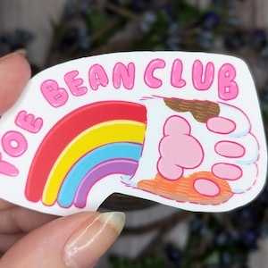 May include: A white sticker with a rainbow and a paw print. The sticker says "Toe Bean Club" in pink bubble letters.
