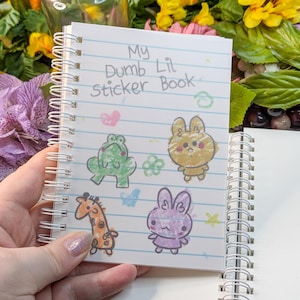 May include: A small spiral-bound notebook with the title "My Dumb Lil Sticker Book" and cartoon animal stickers. The stickers feature a green frog, a yellow bunny, a giraffe, and a purple bunny. The notebook is held in a hand.