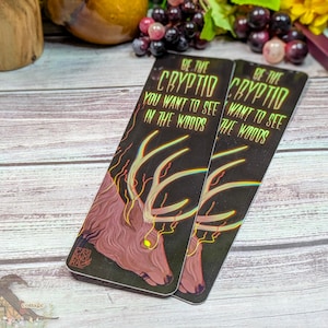 Op de afbeelding: Twee rechthoekige bladwijzers met een donkere achtergrond en een hertenillustratie. De tekst "BE THE CRYPTID YOU WANT TO SEE IN THE WOODS" staat bovenaan. Het hert heeft felgele ogen en een gewei.