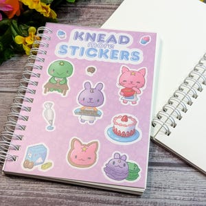 May include: A spiral-bound notebook with a pink cover displaying the text "Knead More Stickers" and cartoon stickers of baked goods and characters. Beside it is an open, blank white notepad. Flowers are visible in the background.