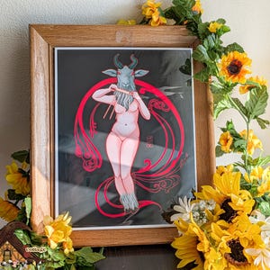 May include: A framed art print featuring a nude figure with a goat head playing a flute, set against a black background with red decorative elements. The artwork is in a wooden frame, surrounded by sunflowers and greenery.