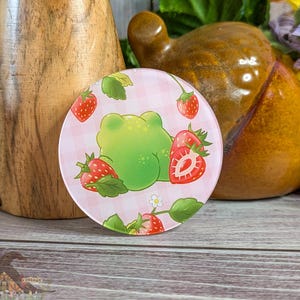 May include: A round, pink coaster with a green frog illustration surrounded by strawberries and leaves. The coaster has a gingham pattern background. The coaster is on a wooden surface.
