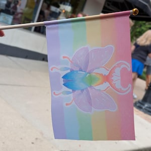 May include: A small rectangular flag attached to a wooden pole featuring a pastel rainbow striped background. Centered on the flag is a stylized illustration of a beetle with translucent light purple wings and a gradient blue and orange body.