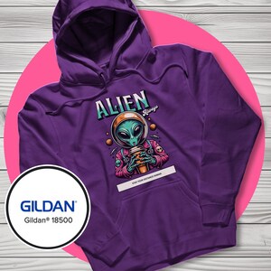 May include: Purple hooded sweatshirt with a graphic of an alien in a space helmet holding a coffee cup. The word "ALIEN" is above the alien, and the text "Stay Your Distance Humans" is below. The sweatshirt has a front pocket and a hood.