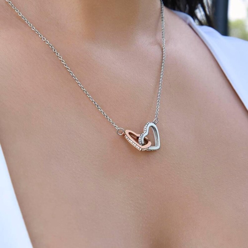 May include: A silver chain necklace with a rose gold and silver heart pendant. The pendant is made of two interlocking hearts, one with a silver outline and the other with a rose gold outline. The hearts are decorated with small, clear stones.