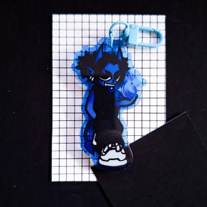 May include: Blue and white character keychain with a blue flame design. The keychain is made of acrylic and has a metal ring.