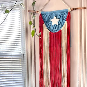 Puede incluir: Un tapiz de pared de macramé con el diseño de la bandera de Puerto Rico. La sección superior es azul claro con una estrella blanca, y la parte inferior tiene rayas verticales rojas y blancas. Está suspendido de un dowel de madera natural con un cordón color crema.