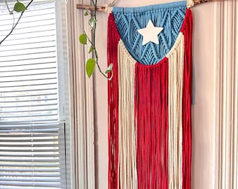 Puede incluir: Un tapiz de pared de macramé con el diseño de la bandera de Puerto Rico. La sección superior es azul claro con una estrella blanca, y la parte inferior tiene rayas verticales rojas y blancas. Está suspendido de un dowel de madera natural con un cordón color crema.