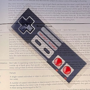 May include: A page from a tabletop role-playing game rulebook, featuring text about removing curses. A clear plastic overlay with a cross-shaped design is placed over the text, with the words "REMOVE CURSE" visible.