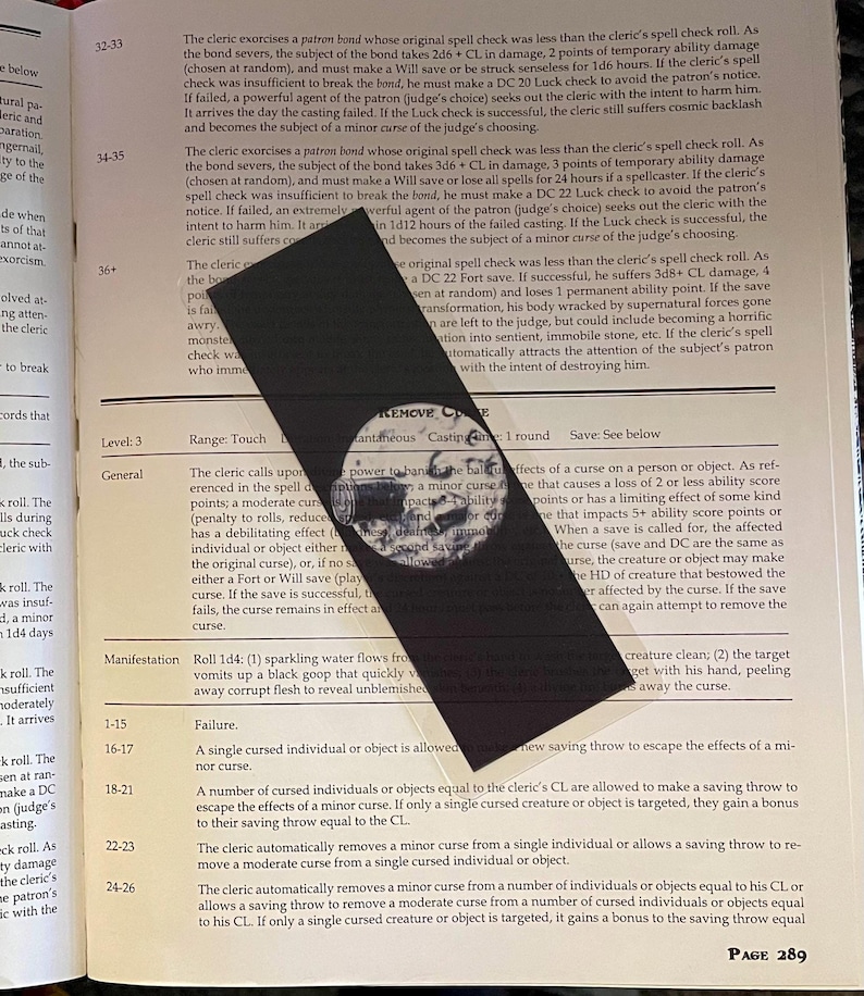 May include: Black bookmark with a skull illustration and the text "REMOVE CURSE" on a page of a book. The bookmark is angled across the page, with text visible on the book.