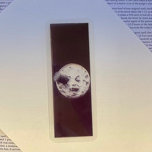 May include: A sticker featuring a black background with a white, smiling moon with a telescope pointed at it. The sticker has a white border and is rectangular in shape.