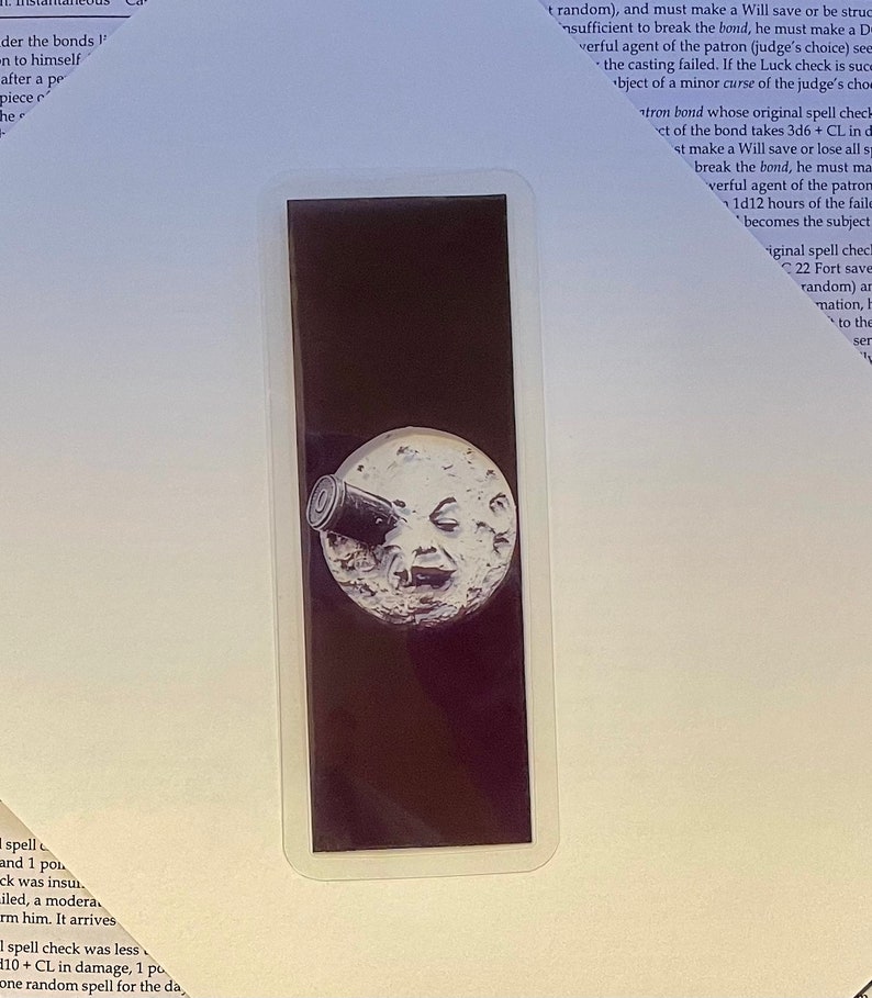 May include: A sticker featuring a black background with a white, smiling moon with a telescope pointed at it. The sticker has a white border and is rectangular in shape.