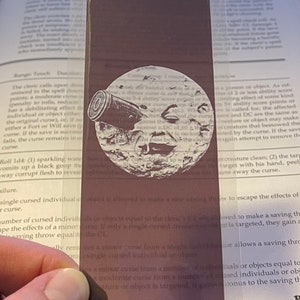May include: A rectangular, dark brown bookmark with a white illustration of a moon with a cannon. The bookmark is made of a transparent material and is placed over a book page.