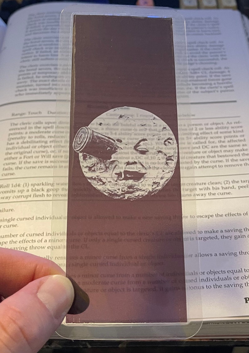 May include: A rectangular, dark brown bookmark with a white illustration of a moon with a cannon. The bookmark is made of a transparent material and is placed over a book page.
