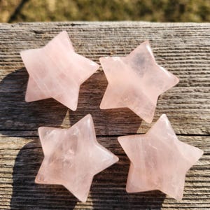 Pode incluir: Quatro pedras polidas em forma de estrela de quartzo rosa. Cada estrela tem cinco pontas e um tom rosa suave. As pedras estão dispostas sobre uma superfície de madeira envelhecida, com sombras.