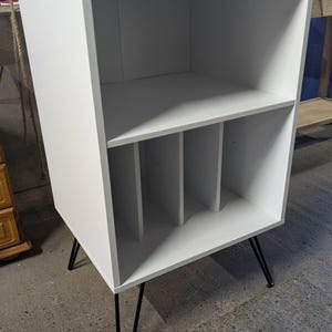 May include: A white storage cabinet with black hairpin legs. The cabinet features an open top shelf and a lower section divided into three compartments. The design is minimalist and functional, suitable for various storage needs.