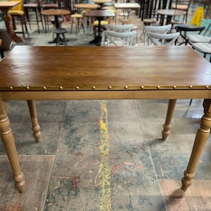 May include: A rectangular wooden table with a light brown finish. The tabletop is adorned with small, evenly spaced brass-colored studs along the edges. The table has four sturdy, turned legs, adding to its classic design. The table is approximately 122 cm long and 76 cm wide.