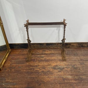 May include: A brown, weathered cast iron stand featuring two vertical supports with ornate, textured detailing connected by a horizontal crossbar at the top and a wider, flat base at the bottom, designed for structural support or display purposes.