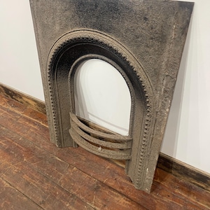 May include: A dark grey cast iron fireplace insert featuring an arched opening with a decorative beaded border and three horizontal bars across the lower section. The metal surface has a textured, weathered finish with visible signs of age.