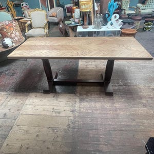 May include: A rectangular dining table featuring a light brown wooden top with a distinct herringbone pattern. The table is supported by two dark brown, thick, cylindrical pedestal legs connected by a horizontal base bar at the bottom.
