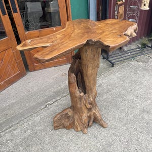 May include: A tall, light brown wooden pedestal table crafted from a single piece of tree trunk. The table features a wide, irregular, flat top surface with natural wood grain patterns and a sturdy, thick base formed by the original root structure.