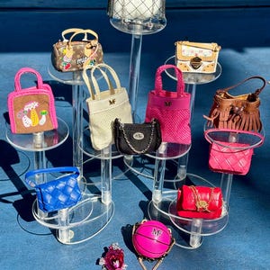 Può includere: Una collezione di mini borse in vari stili e colori, tra cui rosa, blu, marrone e nero. Alcune borse presentano tracolle a catena, mentre altre hanno manici superiori. Le borse sono esposte su supporti trasparenti, mostrando i loro dettagli.