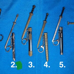 May include: Miniature sword keychains in various designs. The collection includes swords with gold, silver, and black finishes, some with decorative sheaths and key rings. The swords are arranged on a blue surface.