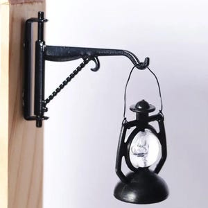 May include: A miniature black lantern with a clear glass globe hangs from a black metal bracket. The bracket is attached to a light-colored wooden surface. The lantern has a rounded base and a handle for hanging.