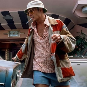 May include: A person wearing a white bucket hat, gold-rimmed sunglasses, a patterned short-sleeved shirt, and a multi-coloured patchwork jacket with red, tan, and grey panels. The person is holding a tall glass of red liquid and has a cigarette.