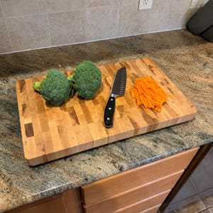 May include: A rectangular wooden cutting board with a block pattern, featuring two heads of green broccoli, a stainless steel santoku knife with a black handle, and a pile of thinly sliced orange carrots arranged on the surface of the board.