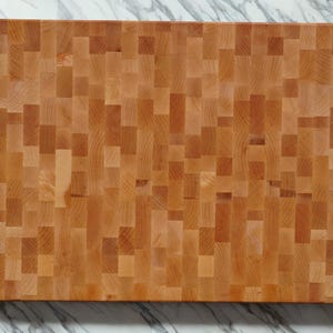 May include: A rectangular end-grain cutting board made of light brown wood with a mosaic-like pattern of small, vertical, and horizontal rectangular blocks. The surface shows natural wood grain variations in shades of tan, beige, and light brown.