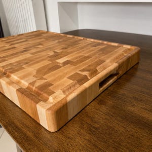 May include: A rectangular end-grain wooden cutting board featuring a light to medium brown mosaic pattern of wood blocks. The board has a smooth surface, rounded edges, and a recessed handle cutout on the side for easy lifting and transport.