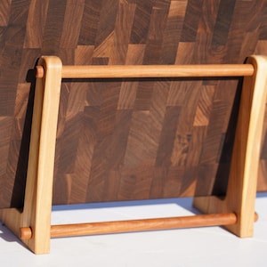 May include: A dark brown butcher block cutting board with a light brown wooden stand. The cutting board is made of end-grain wood, and the stand is made of light-colored wood. The stand has a horizontal bar and two vertical supports.