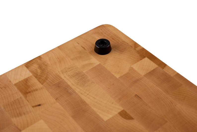 May include: A light brown wooden cutting board with a butcher block pattern made of small rectangular wood pieces. A single small black circular rubber foot is attached to the corner of the board, providing a stable base for the wooden surface.