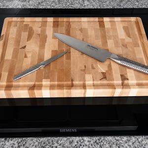 May include: A rectangular wooden cutting board with a light brown block pattern and a grooved edge, featuring a large Global chef knife and a smaller paring knife with metallic dimpled handles resting on the surface of the board.