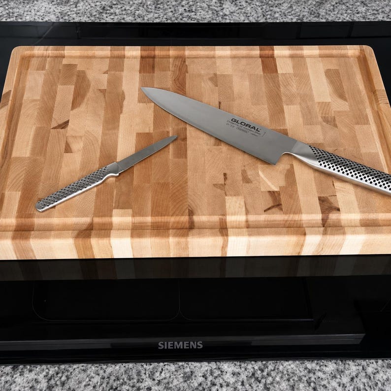 May include: A rectangular wooden cutting board with a light brown block pattern and a grooved edge, featuring a large Global chef knife and a smaller paring knife with metallic dimpled handles resting on the surface of the board.