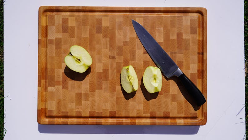 May include: A rectangular wooden cutting board with a light and medium brown checkered pattern, featuring a juice groove along the perimeter. Three pale green apple slices are arranged on the board next to a stainless steel chef's knife.