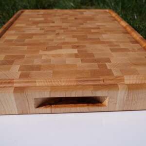 May include: A rectangular end-grain wooden cutting board featuring a light tan and honey-colored mosaic pattern. The board includes a carved juice groove along the perimeter and a recessed handle cutout on the front edge for easy lifting.
